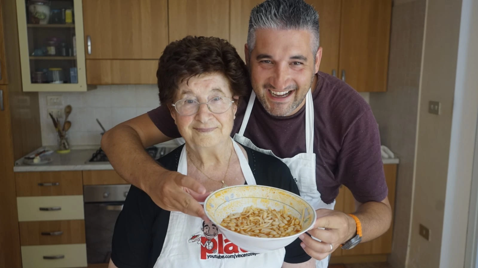 Vincenzo Prosperi in a kitchen with an older woman holding a bowl of food, smiling toward the camera