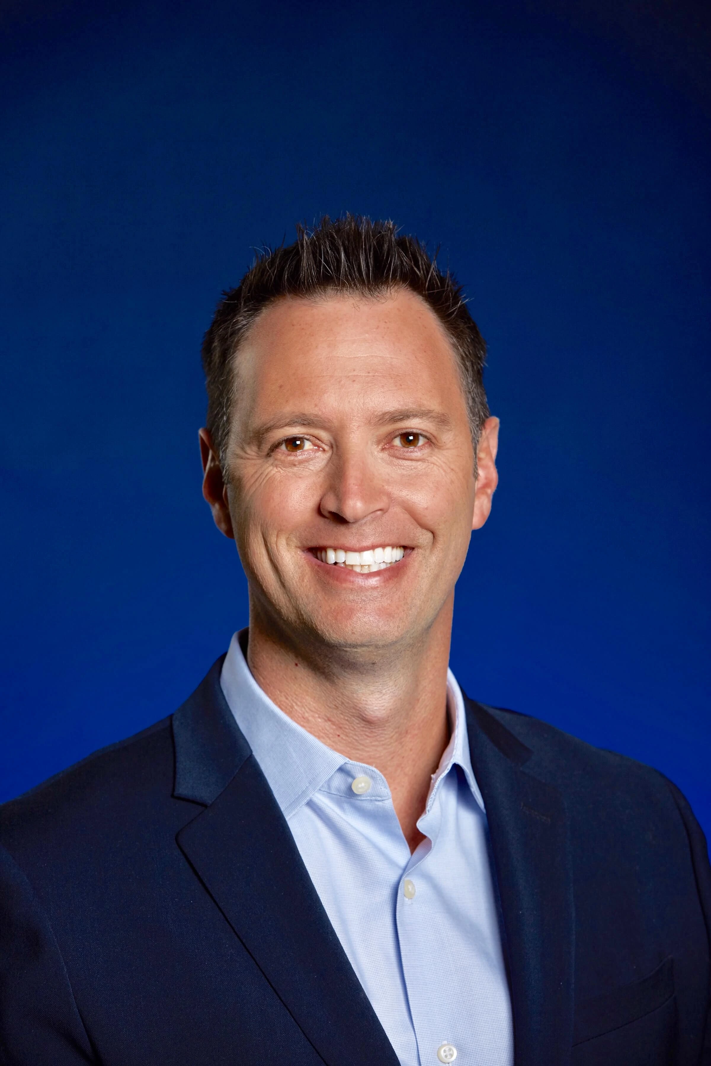 Portrait headshot of Gio Valiante in a dark suit against a blue background