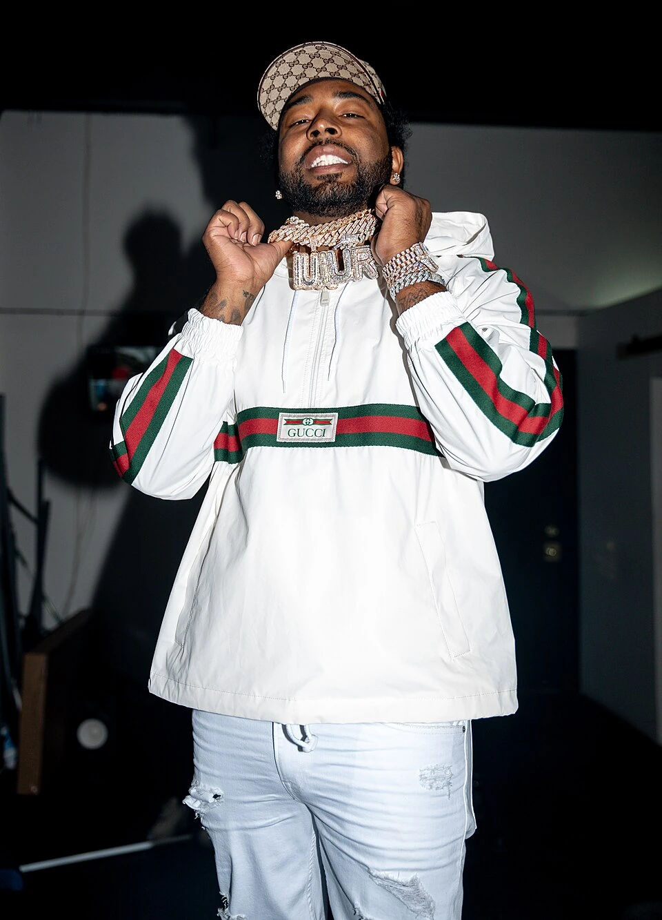 Icewear Vezzo posing indoors wearing a white Gucci tracksuit and cap, holding up his chain.