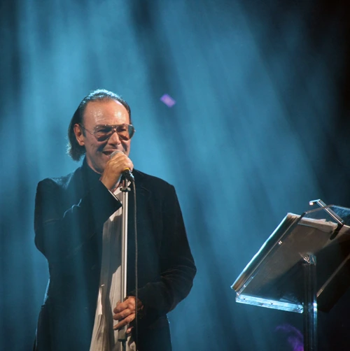 Antonello Venditti performing on stage with a microphone and music stand under blue lighting.
