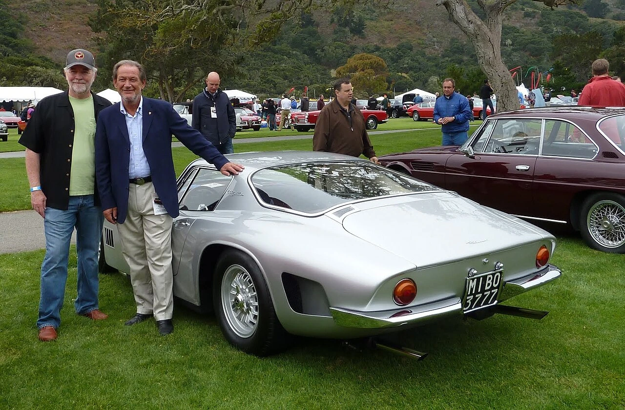Valentino Balboni standing beside a silver Bizzarrini GT 5300 Strada at an outdoor car event