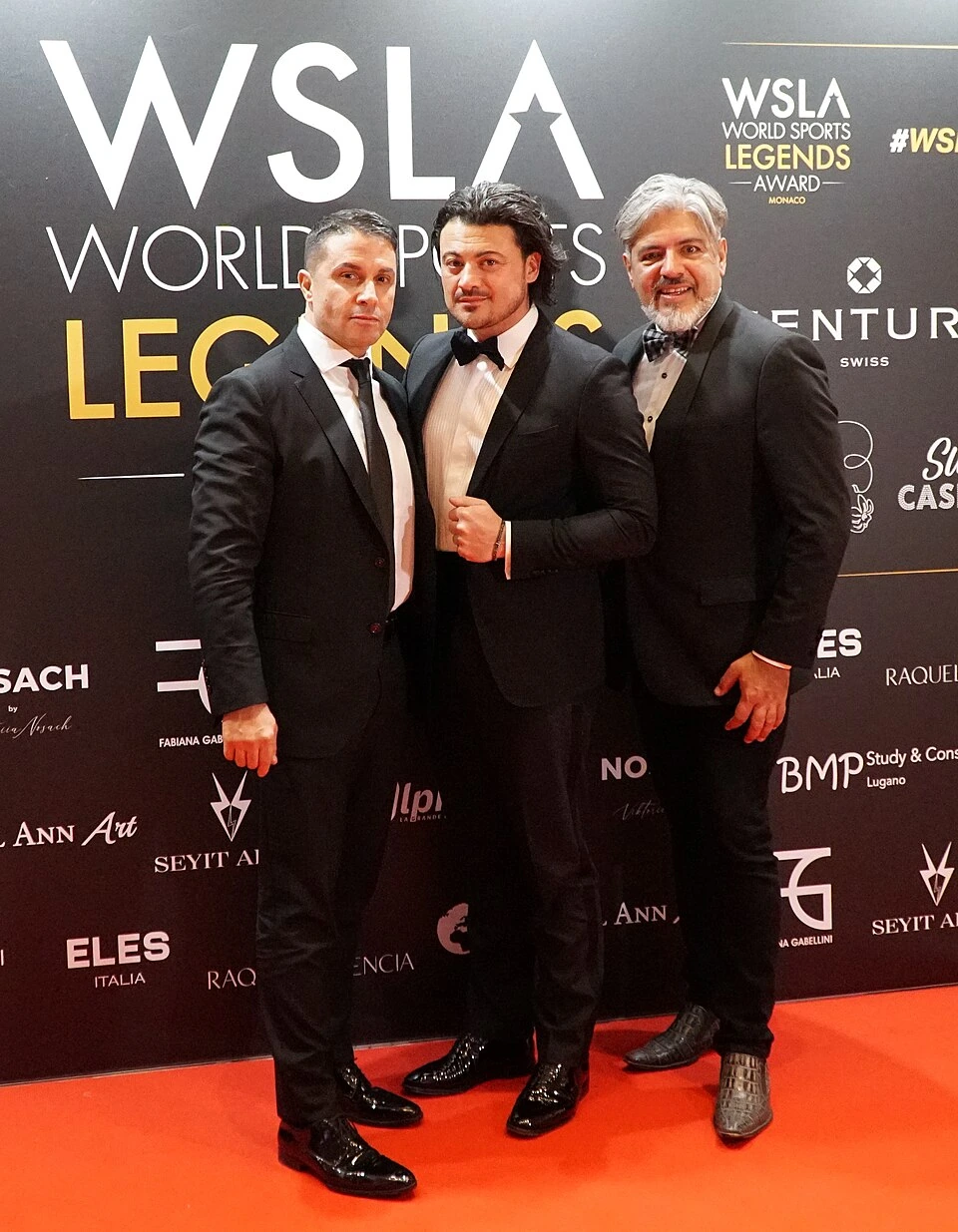 Vittorio Grigolo poses on the red carpet in a tuxedo at a WSLA event with two other men.