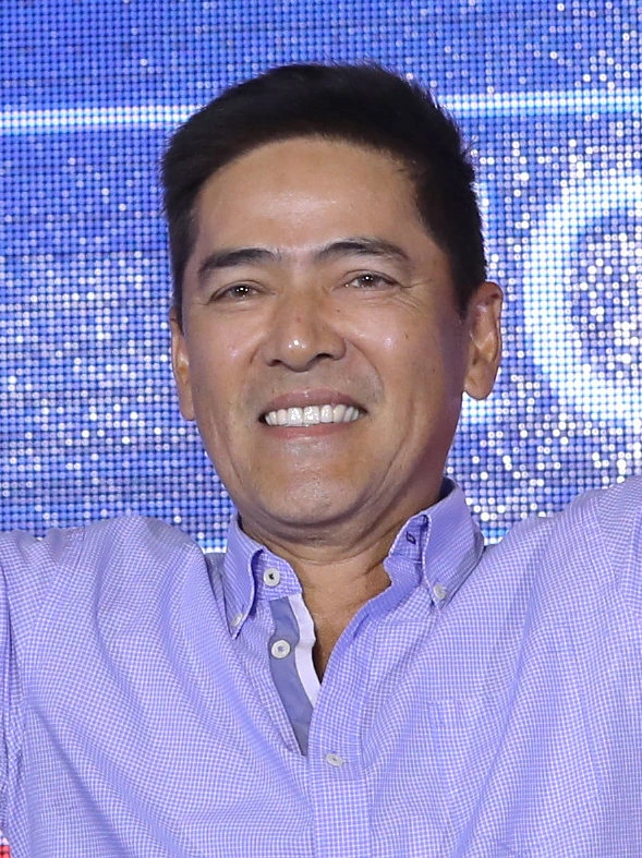 Vic Sotto smiling in a purple shirt against a blue LED backdrop